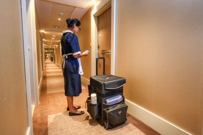 Une femme de chambre dans le couloir d'un hôtel avec une valisette à roulette.